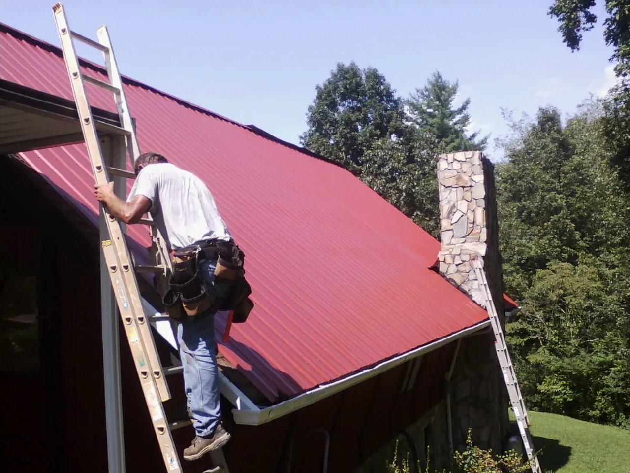 Roofer on a ladder beside a red metal roof and stone chimney — Soffit & Fascia Repair by Roofing Brothers in Marion Station
